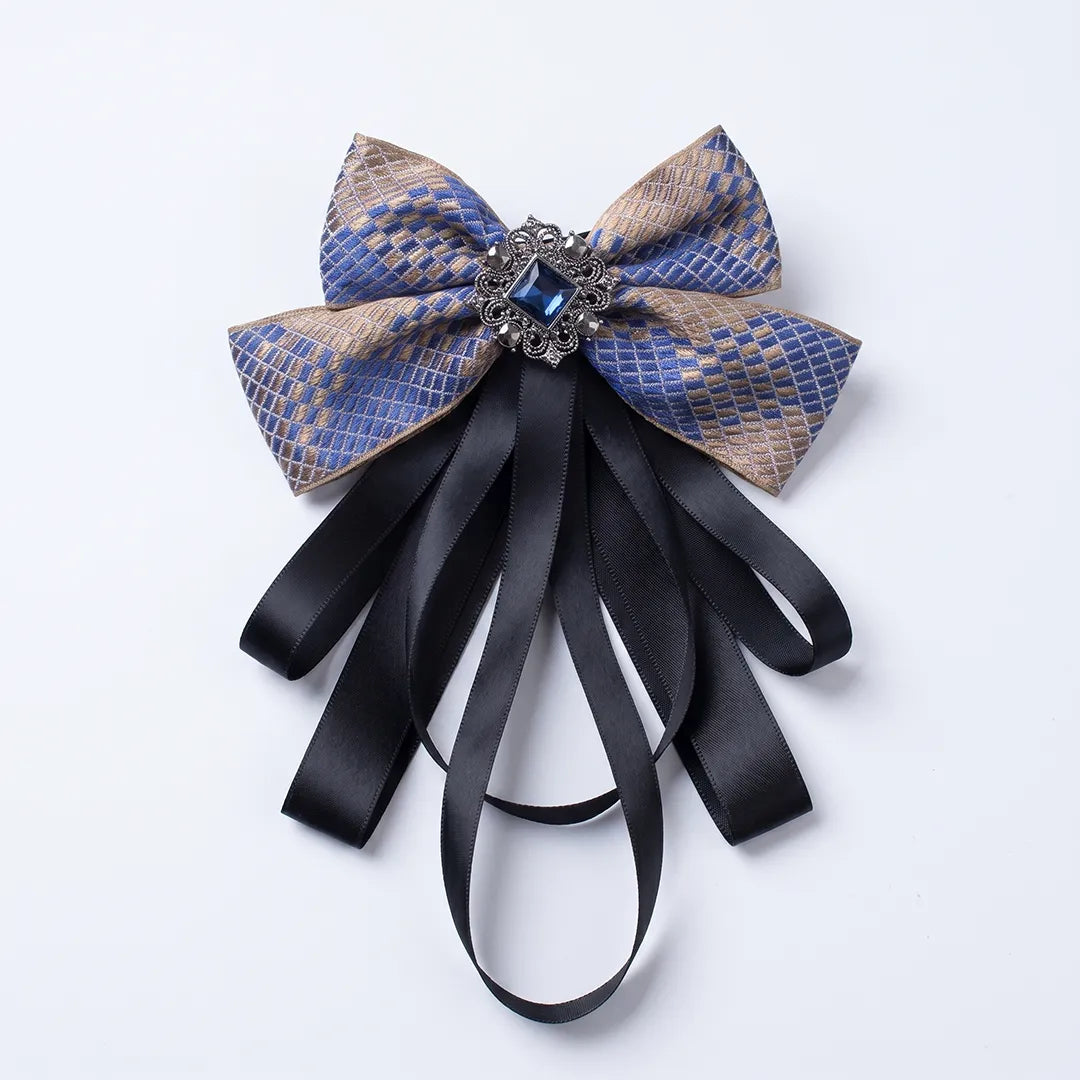 Close-up of a luxurious blue and gold checkered silk bow tie featuring cascading black ribbon tails and an ornate square blue crystal brooch.
