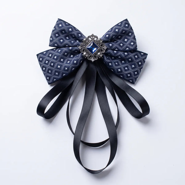 Close-up of a luxurious navy blue geometric silk bow tie featuring cascading black ribbon tails and an ornate square blue crystal brooch.