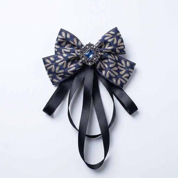Close-up of a luxurious navy blue and gold leaf silk bow tie featuring cascading black ribbon tails and an ornate square blue crystal brooch.