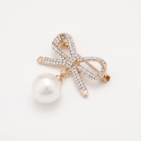 Close-up of a rose gold-tone ribbon bow collar pin encrusted with clear crystals and featuring a large dangling white faux-pearl.