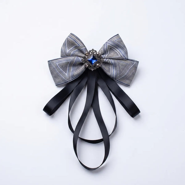 Close-up of a luxurious silver grey silk bow tie featuring cascading black ribbon tails and an ornate square blue crystal brooch.