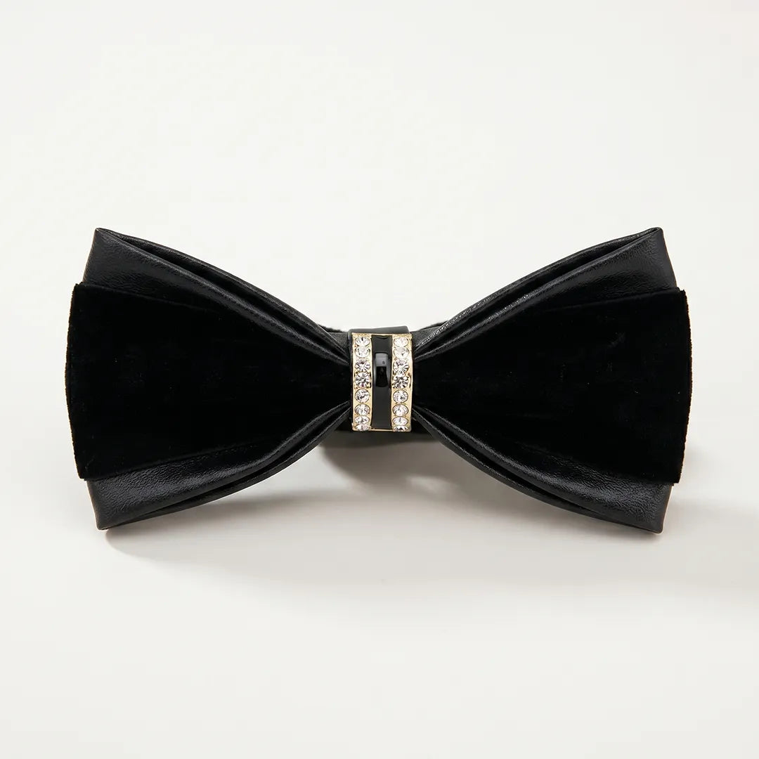 Close-up of a luxurious all-black leather and velvet bow tie featuring a gold metallic ring centerpiece encrusted with clear crystals and black enamel.