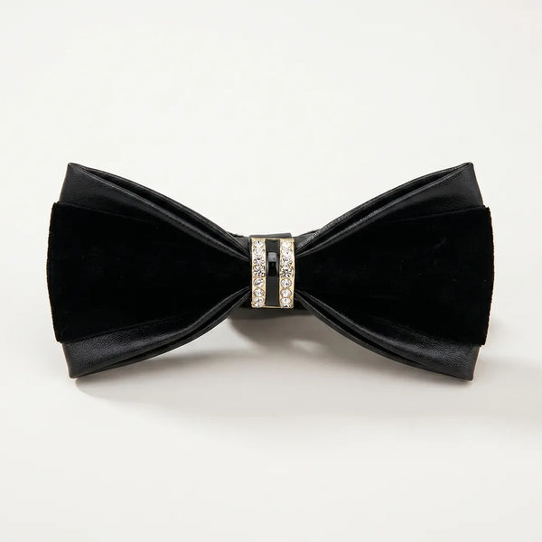 Close-up of a luxurious all-black leather and velvet bow tie featuring a gold metallic ring centerpiece encrusted with clear crystals and black enamel.