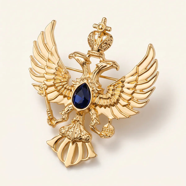 Close-up of a polished gold-tone double-headed eagle crest collar pin featuring a crown, scepter, orb, and a sapphire-blue center stone.