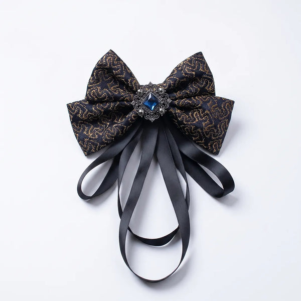 Close-up of a luxurious black and gold baroque silk bow tie featuring cascading black ribbon tails and an ornate square blue crystal brooch.
