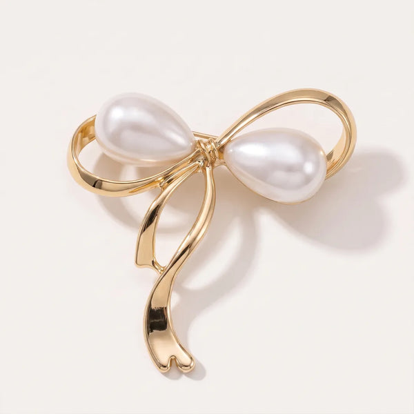 Close-up of a polished gold-tone ribbon bow collar pin featuring two large white teardrop faux-pearls integrated into the design.
