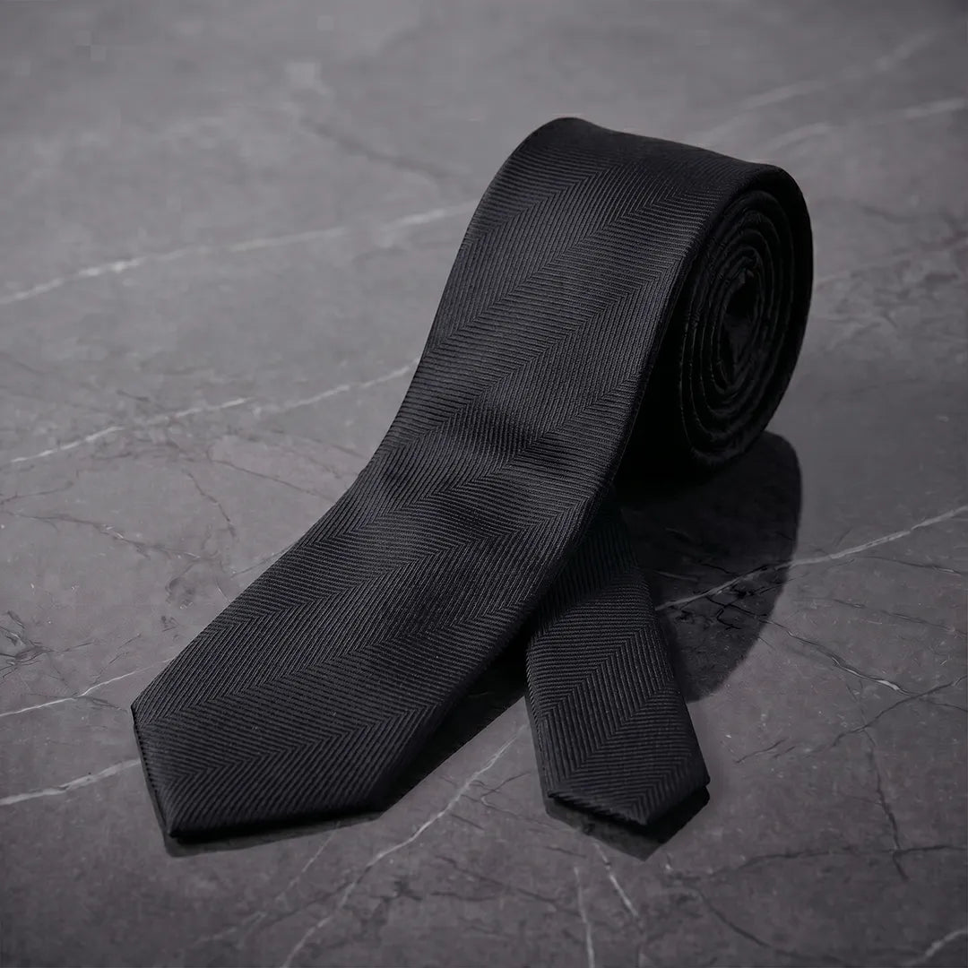 Rolled black herringbone skinny tie styled on a dark marble surface.