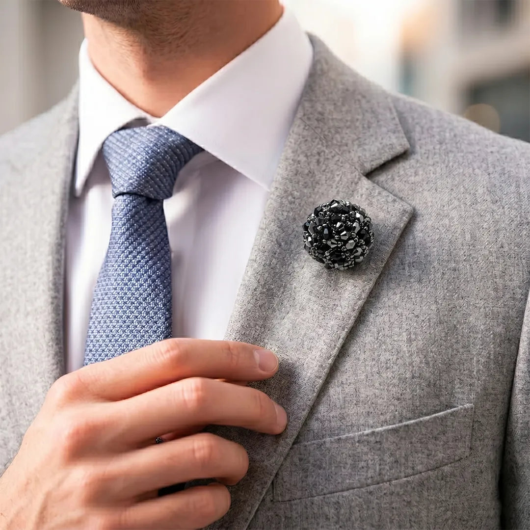 Close-up of a man in a light grey suit and blue tie adjusting his lapel, wearing a black and silver beaded lapel pin.