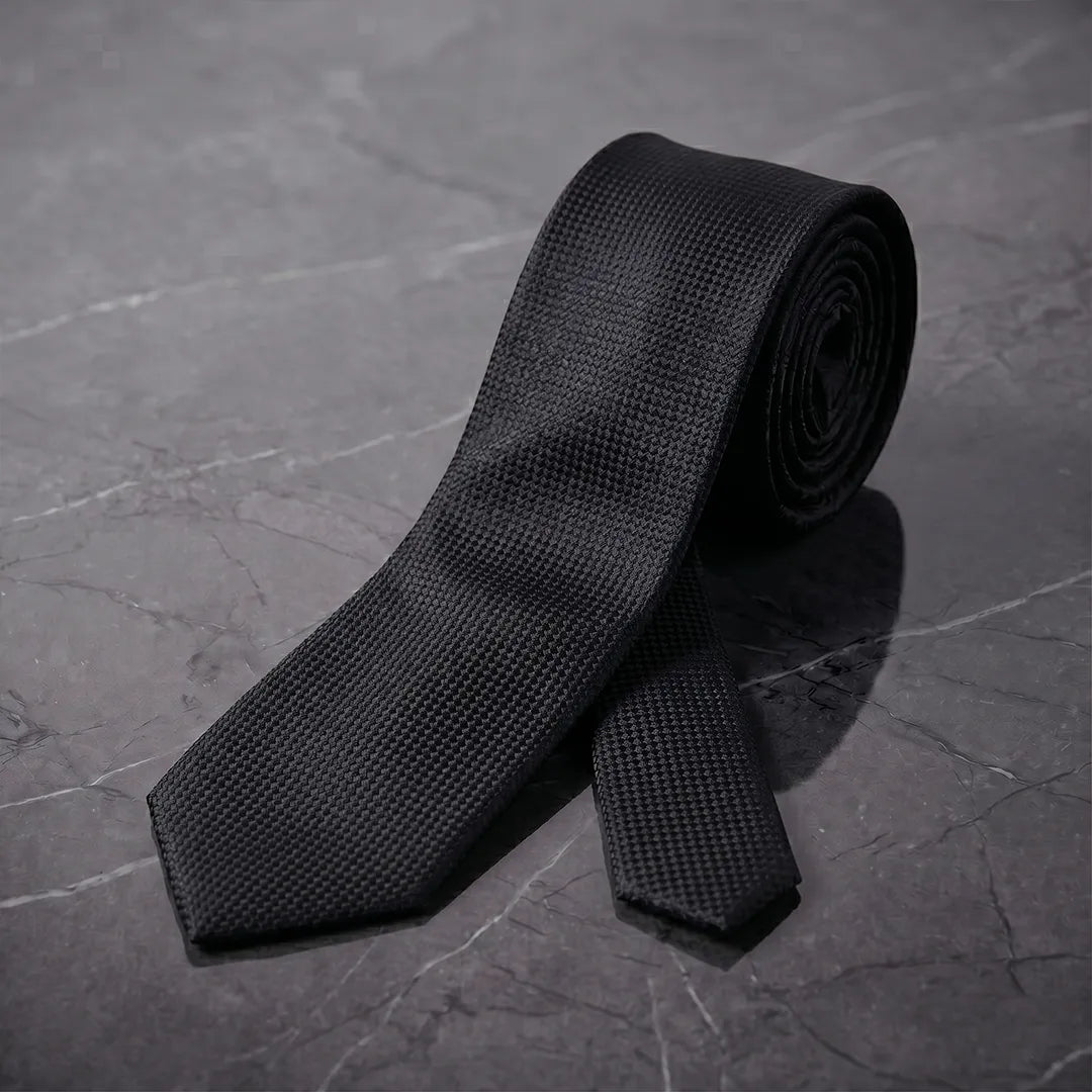 Rolled black woven cotton skinny tie styled on a dark marble surface.