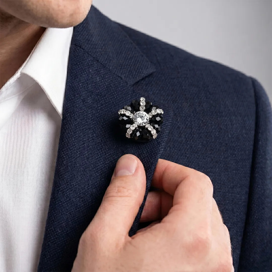 Close-up of a man in a navy suit adjusting his lapel, wearing a black and crystal flower lapel pin with a white dress shirt.