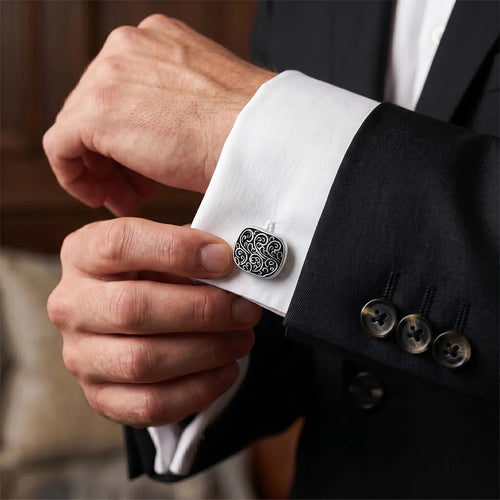 Close-up of a man's hand fastening an antique silver filigree cufflink onto a white French-cuff shirt.