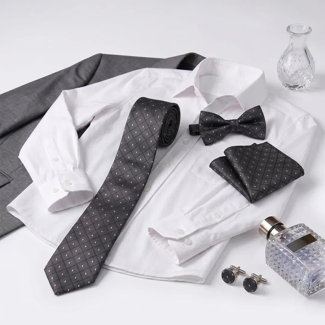 A men's formal accessory set arranged on a white dress shirt, including a dark gray patterned necktie, matching bow tie, pocket square, and cufflinks, with a gray suit jacket and a small glass vase in the background.