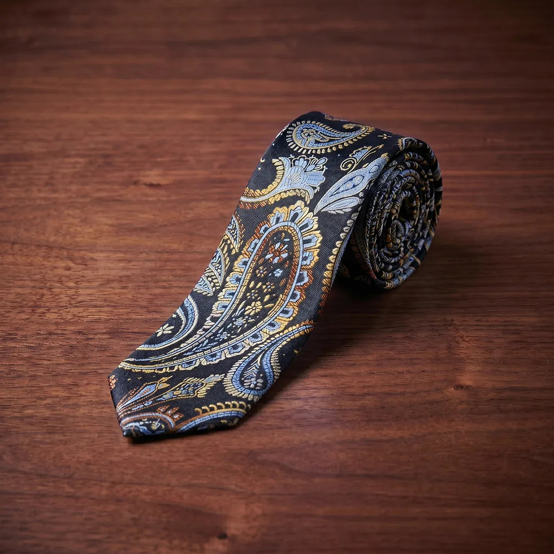 A rolled necktie with a detailed paisley pattern in blue, gold, and white on a black base, displayed on a dark brown wooden surface.