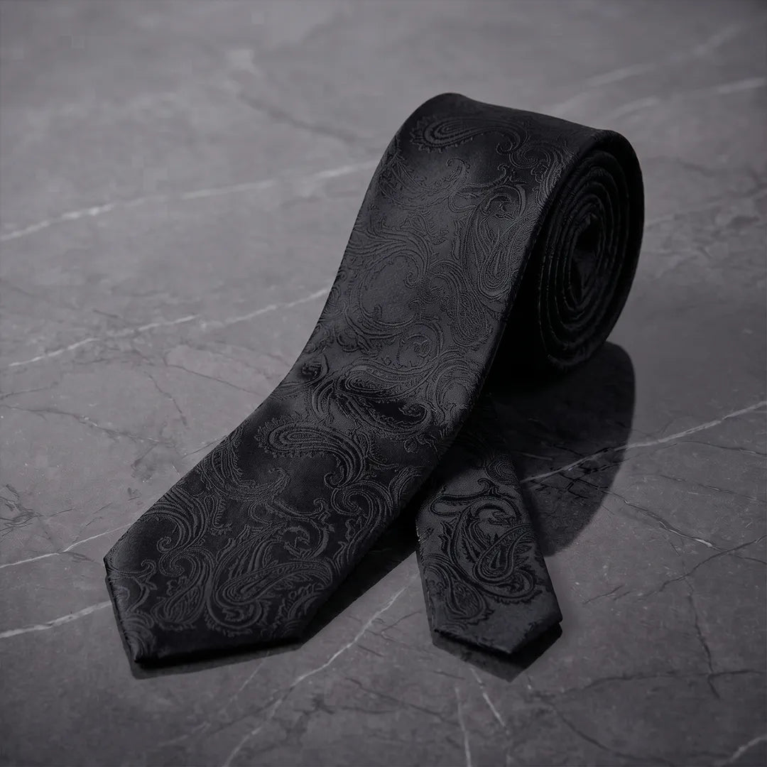 A black silk necktie with a subtle embossed paisley pattern, displayed on a dark gray marble surface.