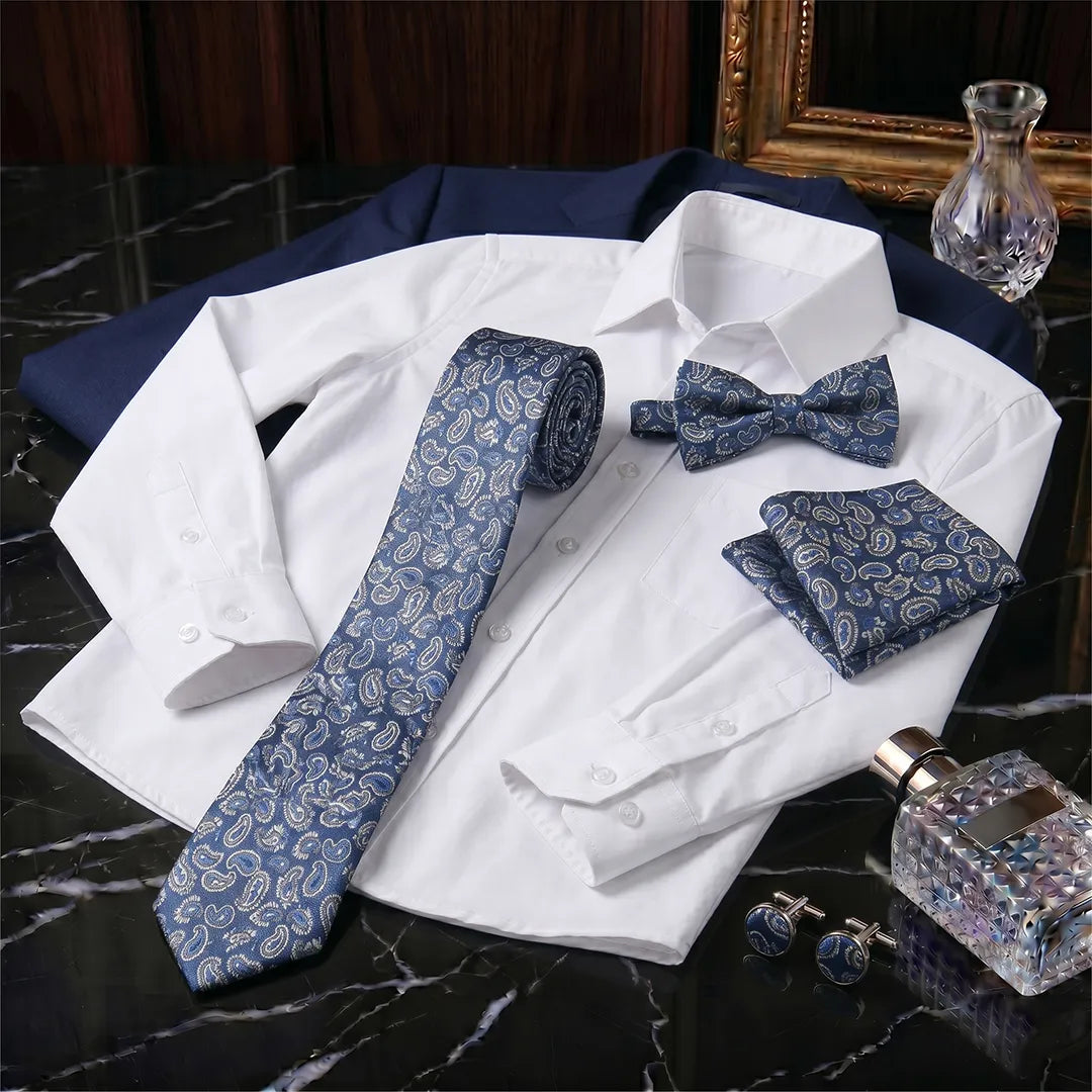A men's formal accessory set arranged on a white dress shirt, including a blue paisley necktie, matching bow tie, pocket square, cufflinks, and a folded navy blazer. The set is styled on a black marble surface with a perfume bottle and framed mirror in the background.