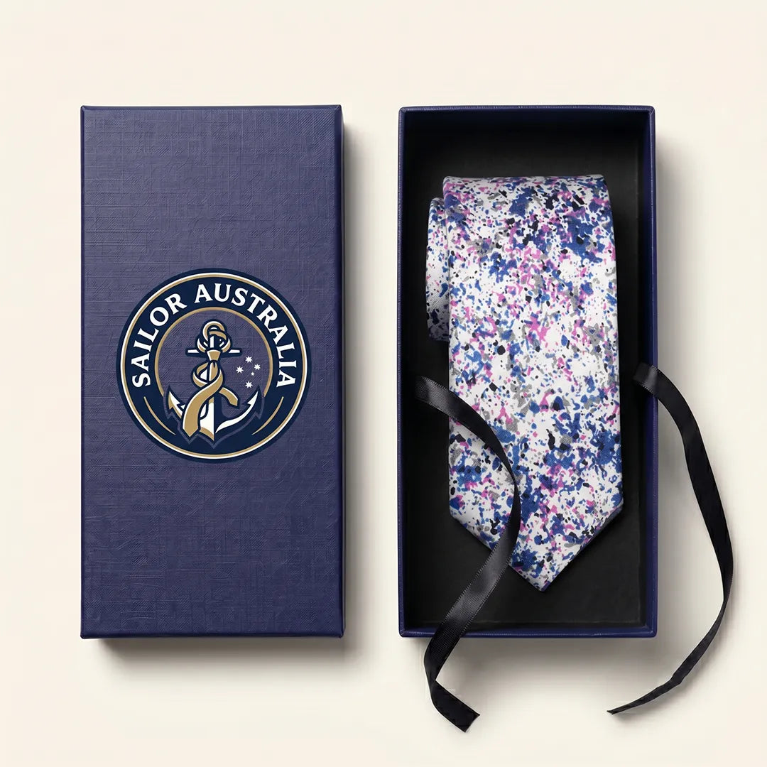 A patterned necktie displayed inside an open dark blue presentation box next to its matching lid, which features a circular 'SAILOR AUSTRALIA' emblem. The tie has a dense floral or abstract print in purple, pink, and blue on a white base, with a black ribbon tied around it.